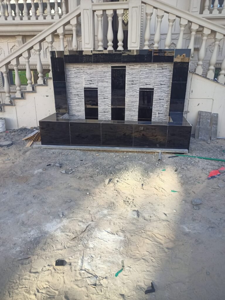 شلال صناعي مع نافورة مائية وحديقة مزينة بأحواض زهور وجلسة خارجية حديثة.